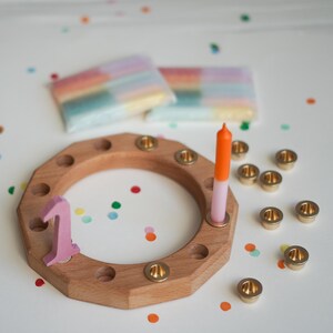 May include: Wooden birthday candle holder with brass candle cups, a pink number one, and a two-tone orange and pink candle. Two packages of rainbow-colored candles are in the background. Colorful confetti dots the white surface.