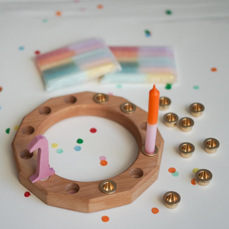 May include: Wooden birthday candle holder with brass candle cups, a pink number one, and a two-tone orange and pink candle. Two packages of rainbow-colored candles are in the background. Colorful confetti dots the white surface.