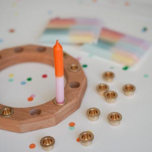 May include: A wooden candle holder with brass inserts, holding an orange and pink candle. Several brass candle cups and colorful confetti are scattered around the holder. Two blurred, colorful paper items are in the background.