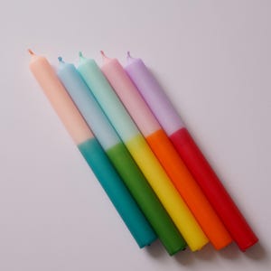 Könnte beinhalten: Sechs bunte Kerzen mit einem zweifarbigen Design. Die obere Hälfte jeder Kerze ist in Pastellfarben gehalten, die untere Hälfte in einer leuchtenden Farbe. Die Farben sind Lavendel, Rosa, Hellblau, Mintgrün, Grün und Gelb.