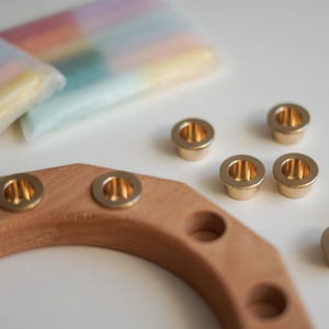 May include: A wooden candle holder with brass-colored metal inserts. Several loose metal inserts are scattered around the holder. Pastel-colored candles are visible in the background, suggesting a home decor theme.