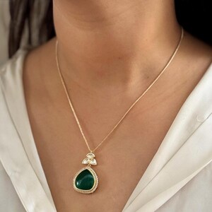 Puede incluir: Un collar de cadena de oro con un colgante en forma de l&aacute;grima. El colgante es verde y tiene un engaste de piedras transparentes alrededor del borde. El colgante est&aacute; acentuado con tres piedras transparentes m&aacute;s peque&ntilde;as.