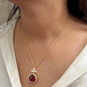 Puede incluir: Un collar de cadena dorada con un colgante rojo en forma de l&aacute;grima. El colgante est&aacute; rodeado de piedras transparentes y tiene un borde dorado.