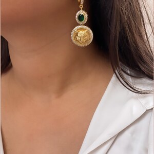May include: Gold hoop earrings with a green gemstone and a circular pendant featuring a detailed design. The pendant is adorned with clear rhinestones.