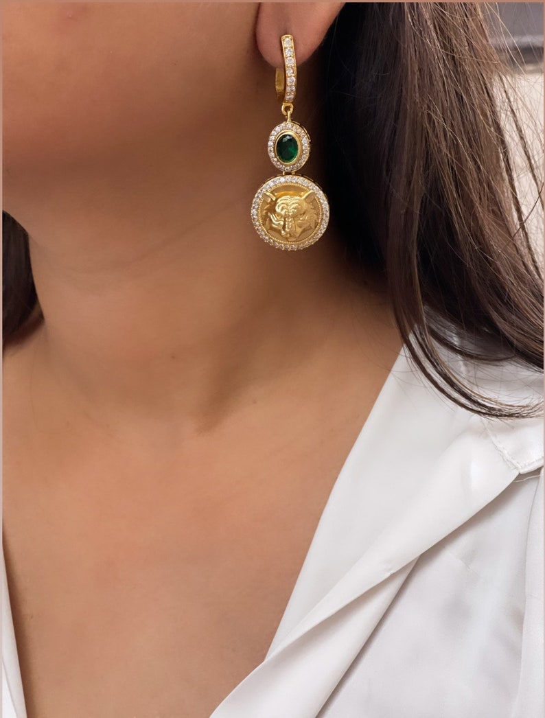 May include: Gold hoop earrings with a green gemstone and a circular pendant featuring a detailed design. The pendant is adorned with clear rhinestones.