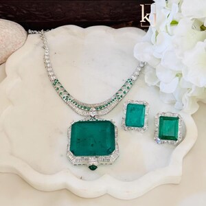 May include: A luxurious jewelry set featuring a necklace and matching earrings. The set includes a large emerald-cut green gemstone pendant and matching earrings, all set in a silver-colored metal with clear accent stones. The necklace has a matching design.
