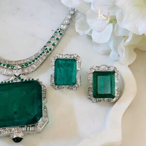 May include: A luxurious jewelry set featuring a necklace, earrings, and a pendant. The pieces showcase large, rectangular emerald-cut green gemstones surrounded by smaller, clear stones. The jewelry is set on a white surface with a floral accent.