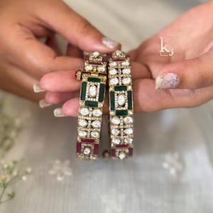 Peut inclure: Deux bracelets dorés aux détails complexes. Chaque bracelet présente un motif rectangulaire vert et rubis, entouré de pierres claires et scintillantes. Les bracelets sont présentés sur un fond doux et clair.