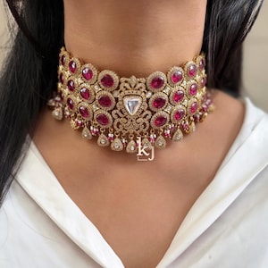May include: A gold choker necklace featuring pink and white gemstones. The necklace has a delicate design with intricate details. The necklace is adorned with dangling charms.
