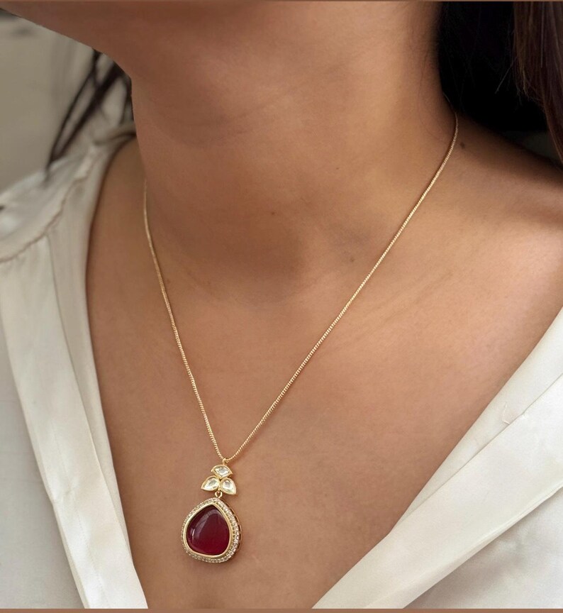 Puede incluir: Un collar de cadena dorada con un colgante rojo en forma de l&aacute;grima. El colgante est&aacute; rodeado de piedras transparentes y tiene un borde dorado.