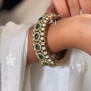 May include: Gold-toned bangle bracelet adorned with emerald-green and clear gemstones. The bracelet features a repeating pattern of emeralds and clear stones, set in a detailed, ornate design. The bracelet is shown on a wrist.