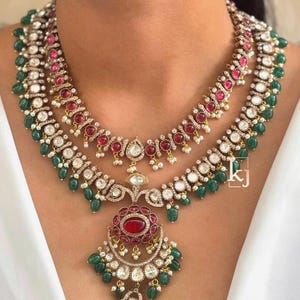May include: A multi-layered necklace featuring red and white gemstones, accented with green beads. The necklace has a large pendant with a central red stone and intricate detailing. The jewelry is set against a white background.