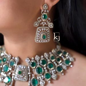 May include: A jewelry set featuring a necklace and a dangling earring with emerald green oval stones and clear white crystals set in a silver-toned metal frame. The earring has a square base with a central green stone. The text kj is visible.