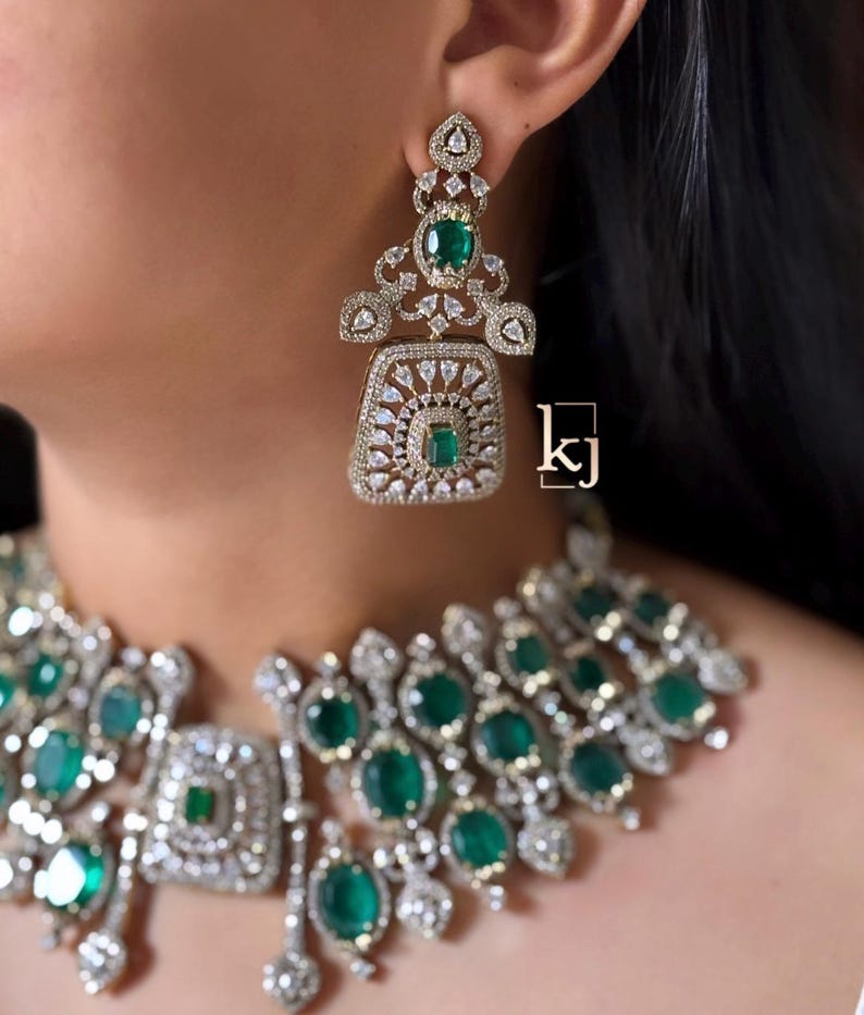 May include: A jewelry set featuring a necklace and a dangling earring with emerald green oval stones and clear white crystals set in a silver-toned metal frame. The earring has a square base with a central green stone. The text kj is visible.