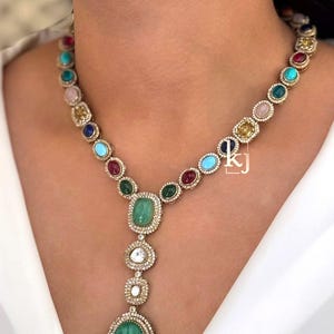 May include: A gold necklace with a variety of colorful gemstones, including emeralds, rubies, and sapphires. The necklace features a pendant with a large green emerald and smaller gemstones. The necklace is set with small, clear stones.