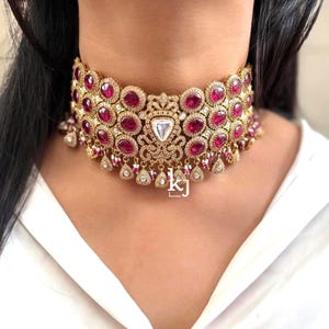 Könnte beinhalten: Ein goldfarbenes Choker-Halsband mit rubinroten Edelsteinen und klaren Kristallen. Die Halskette hat einen zentralen Anhänger mit einem großen, dreieckigen, klaren Stein und aufwendigen Filigranarbeiten. Getragen auf einer weißen Bluse.
