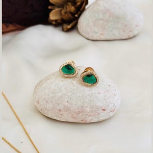 Puede incluir: Un par de pendientes de oro con detalles de piedras preciosas verdes. Los pendientes est&aacute;n colocados sobre una superficie de piedra blanca.