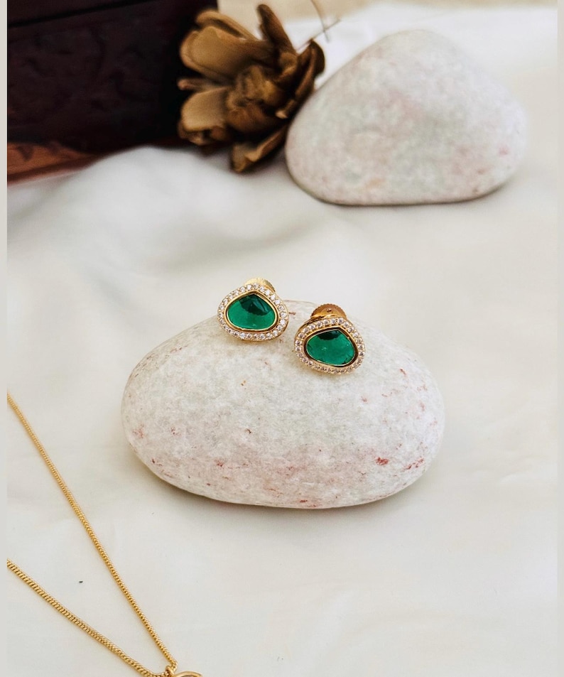 Puede incluir: Un par de pendientes de oro con detalles de piedras preciosas verdes. Los pendientes est&aacute;n colocados sobre una superficie de piedra blanca.