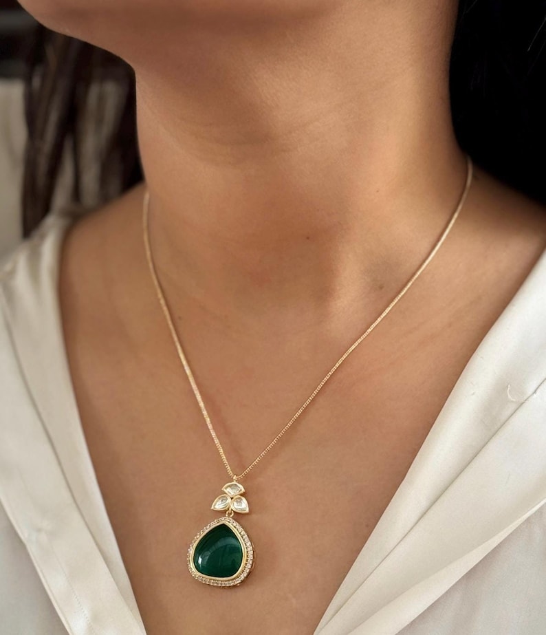Puede incluir: Un collar de cadena de oro con un colgante en forma de l&aacute;grima. El colgante es verde y tiene un engaste de piedras transparentes alrededor del borde. El colgante est&aacute; acentuado con tres piedras transparentes m&aacute;s peque&ntilde;as.