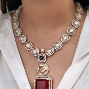 May include: A gold necklace featuring a large red gemstone pendant. The necklace is crafted from small round stones and boasts a detailed design. The pendant is encircled by smaller stones and has a gold design.