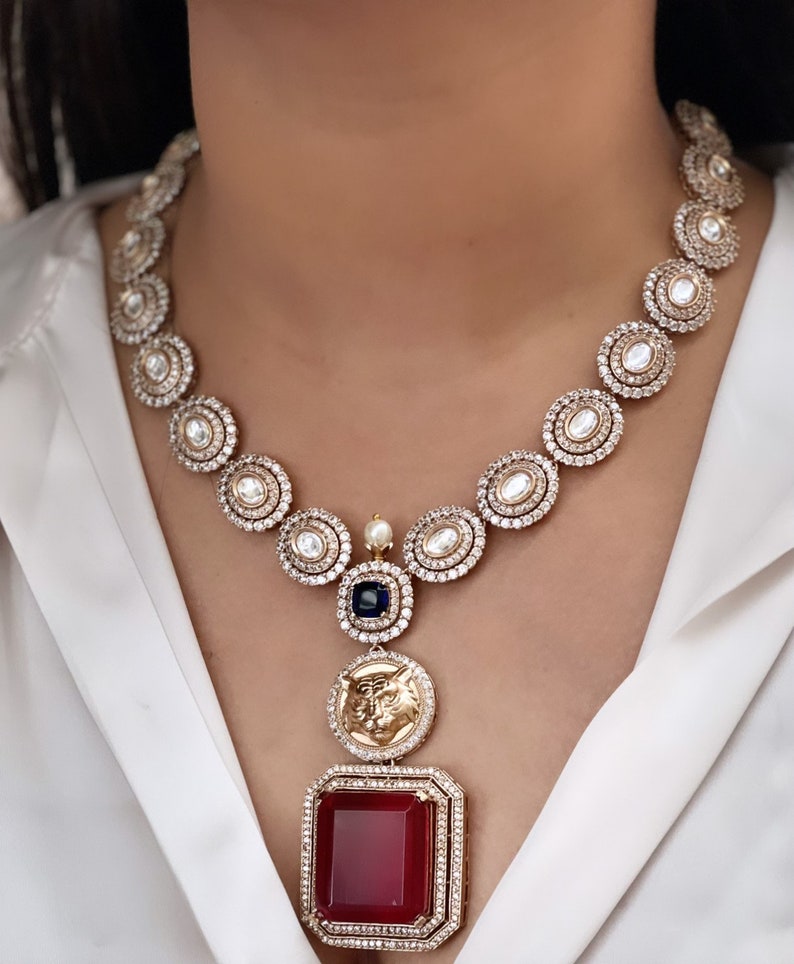 May include: A gold necklace featuring a large red gemstone pendant. The necklace is crafted from small round stones and boasts a detailed design. The pendant is encircled by smaller stones and has a gold design.