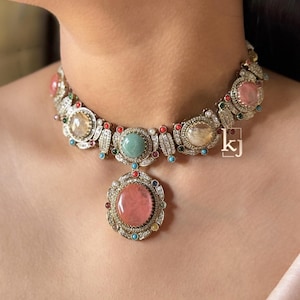 May include: A multi-gemstone necklace with a large pink central stone pendant. The necklace features various colored stones, including pink, green, yellow, and blue, set in a silver-toned metal. The design includes small clear stones and intricate detailing.