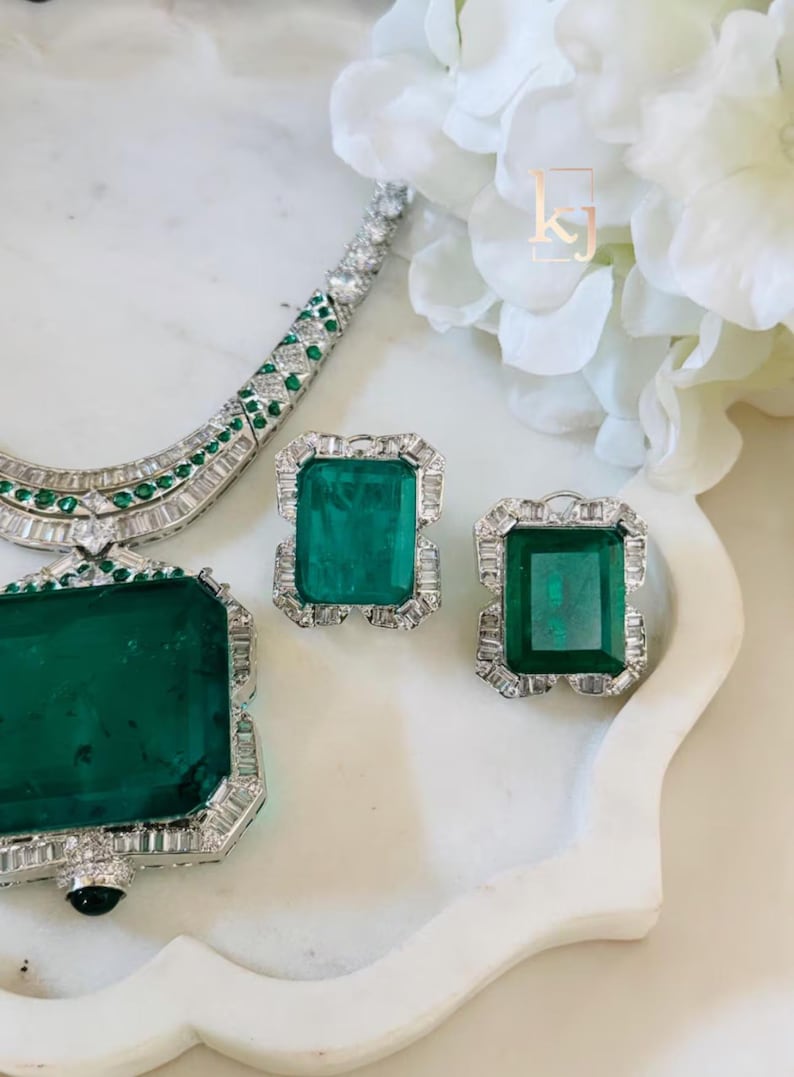 May include: A luxurious jewelry set featuring a necklace, earrings, and a pendant. The pieces showcase large, rectangular emerald-cut green gemstones surrounded by smaller, clear stones, set in a silver or white metal. The jewelry is displayed on a white surface.