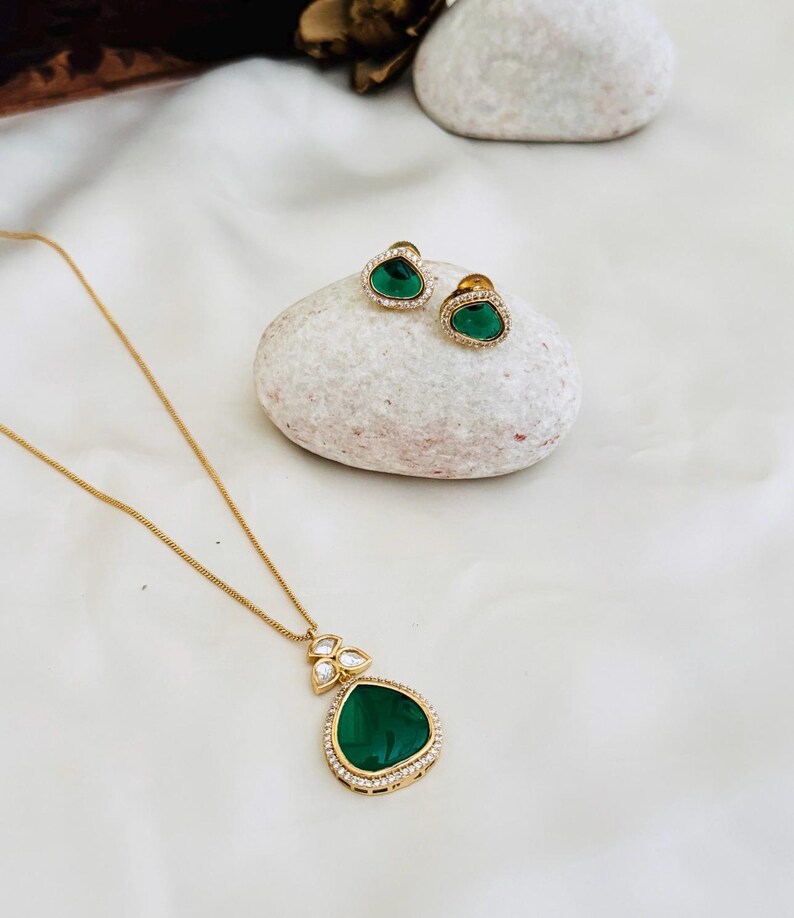 Puede incluir: Un collar de oro con un colgante de piedra preciosa verde y pendientes a juego. El colgante est&aacute; rodeado de piedras preciosas transparentes m&aacute;s peque&ntilde;as.