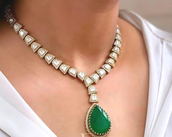 Op de afbeelding: Een goudkleurige ketting met driehoekige, met diamanten bezette schakels. De ketting heeft een druppelvormige, smaragdgroene hanger, eveneens bezet met diamanten. De ketting wordt gedragen op een wit overhemd.