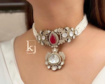 Op de afbeelding: Een goudkleurige choker ketting met een centrale rode edelsteen en heldere, ronde accentstenen. De ketting heeft een bloemenontwerp en een witte kralenstreng. De hanger is een grote, heldere, peervormige steen.