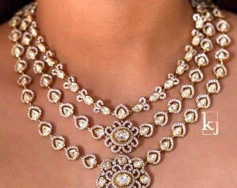 May include: A three-strand gold necklace featuring teardrop-shaped stones surrounded by small white pavé crystals. The two lower strands feature central floral-shaped pendants with additional dangling teardrop accents. The letters kj are visible.