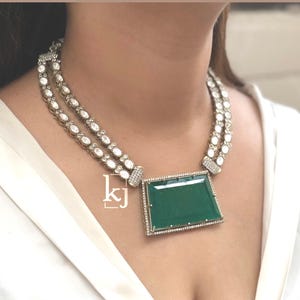 May include: A silver-toned necklace with two strands of white gemstones and a large, emerald-green rectangular pendant. The pendant is framed with small, clear gemstones. The necklace is worn on a white top.