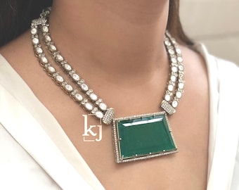 Op de afbeelding: Een zilverkleurige ketting met twee strengen witte edelstenen en een grote, rechthoekige, smaragdgroene hanger. De hanger is omlijst met kleine, heldere edelstenen. De ketting wordt gedragen over een witte top.