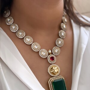 May include: A gold necklace with a large green gemstone pendant. The necklace is made of round, clear stones and has a smaller red gemstone at the top of the pendant.