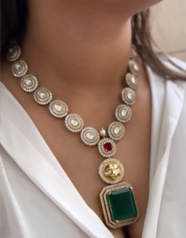 May include: A gold necklace with a large green gemstone pendant. The necklace is made of round, clear stones and has a smaller red gemstone at the top of the pendant.