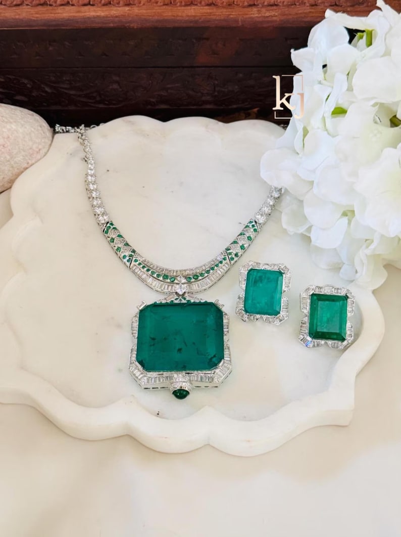 May include: A luxurious jewelry set featuring a necklace and matching earrings. The set includes a large emerald-cut green gemstone pendant and matching earrings, all set in a silver-colored metal with clear accent stones. The necklace is approximately 18 inches long.