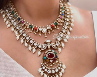 May include: A two-strand gold-toned necklace with colorful gemstones and white pearls. The necklace features a large pendant with a central red stone and a crescent-shaped design. The jewelry is worn on a white garment.
