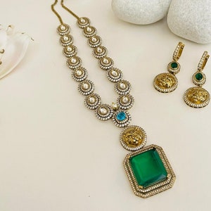May include: A gold necklace with a large green gemstone pendant and smaller blue gemstones. The necklace is made of round, gold-toned beads with white stones. The pendant features a lion's head design. The necklace is paired with matching earrings with green gemstones and lion's head designs.
