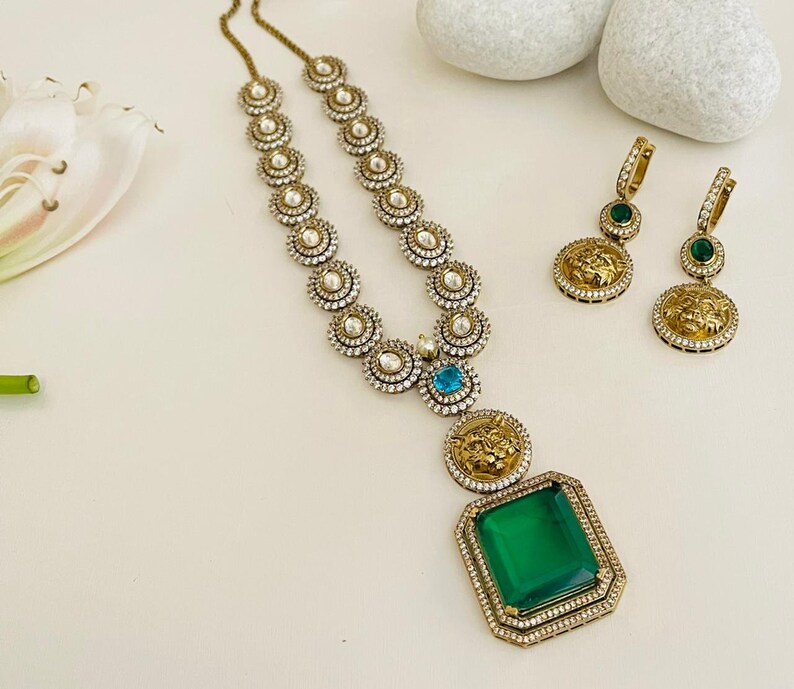 May include: A gold necklace with a large green gemstone pendant and smaller blue gemstones. The necklace is made of round, gold-toned beads with white stones. The pendant features a lion's head design. The necklace is paired with matching earrings with green gemstones and lion's head designs.