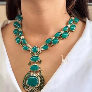 May include: A gold-toned necklace featuring multiple oval-shaped green cabochon stones, accented with small clear crystals and smaller green stones. The design includes a large central pendant drop and a logo with the letters kj.
