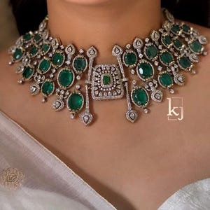 May include: A wide choker necklace featuring multiple oval-shaped green gemstones set in silver-toned metal with intricate clear stone detailing. A central rectangular pendant piece hangs from the collar, and the letters kj are visible.