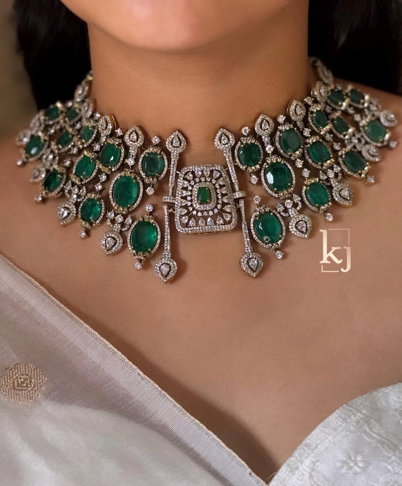 May include: A wide choker necklace featuring multiple oval-shaped green gemstones set in silver-toned metal with intricate clear stone detailing. A central rectangular pendant piece hangs from the collar, and the letters kj are visible.