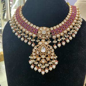 Peut inclure: Un collier doré avec un pendentif central et un design ras du cou. Le collier présente des pierres précieuses rouges et des perles blanches. Le pendentif a une grosse pierre centrale en forme de goutte d'eau et est entouré de petites pierres et de perles.