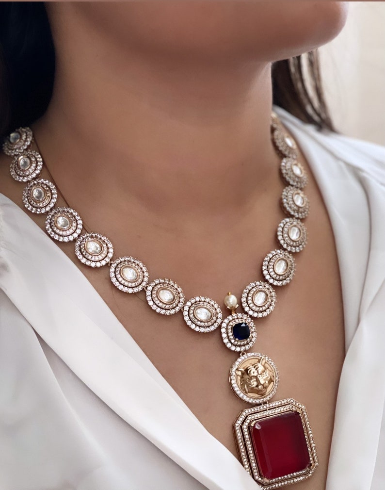 May include: A gold necklace with a red stone pendant. The necklace is made of round, white stones set in gold. The pendant is a square with a red stone in the center and a gold lion head design above it. The necklace is on a white background.