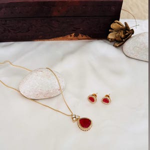 Puede incluir: Un collar de oro con un colgante de l&aacute;grima roja y pendientes a juego. El colgante est&aacute; rodeado de peque&ntilde;as piedras blancas.