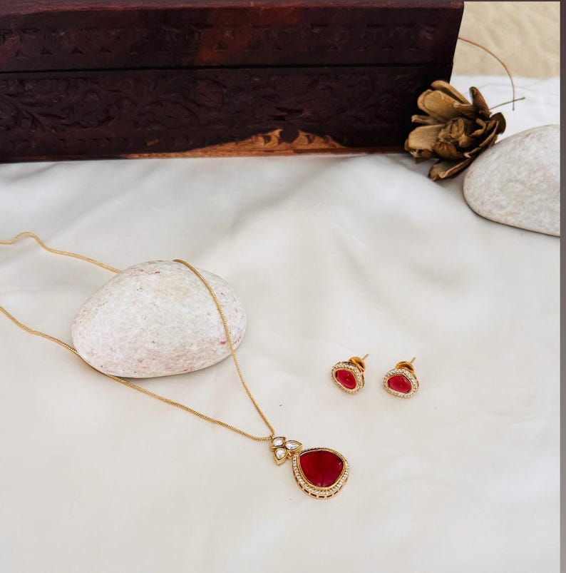 Puede incluir: Un collar de oro con un colgante de l&aacute;grima roja y pendientes a juego. El colgante est&aacute; rodeado de peque&ntilde;as piedras blancas.