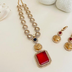 May include: A gold necklace with a red gemstone pendant and a lion head design. The necklace is made of small round beads and has a black gemstone accent. The earrings are gold with red gemstones and lion head designs.