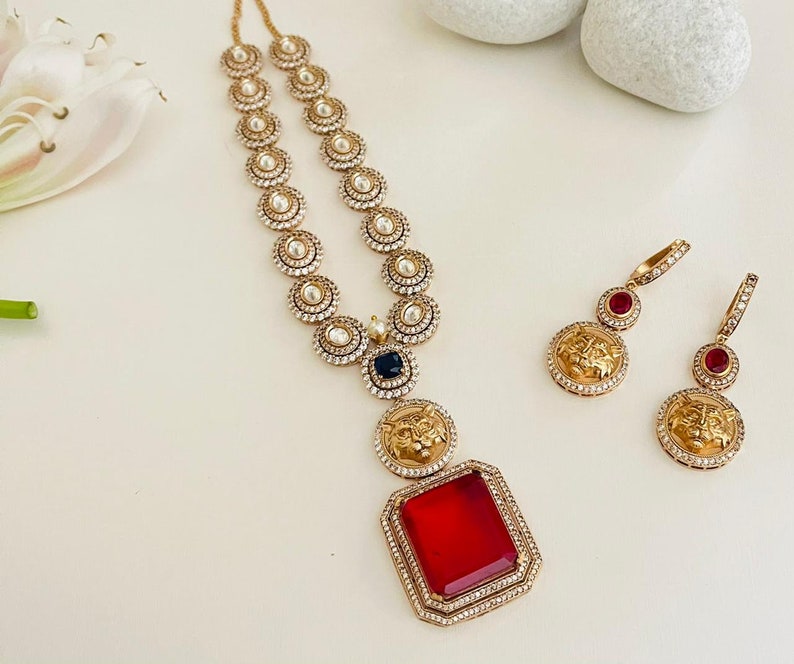 May include: A gold necklace with a red gemstone pendant and a lion head design. The necklace is made of small round beads and has a black gemstone accent. The earrings are gold with red gemstones and lion head designs.