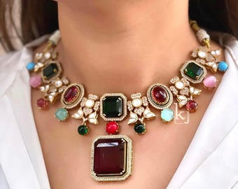 May include: A statement necklace featuring large, colorful gemstones in shades of green, red, and blue. The necklace has a gold-toned setting with intricate detailing and smaller accent stones. The design includes floral elements and a large, rectangular pendant.