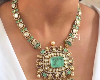May include: A gold-toned necklace featuring a large, rectangular emerald pendant surrounded by diamonds. The necklace has a floral design with emerald and diamond accents, and pearl drops. The necklace is worn around the neck.